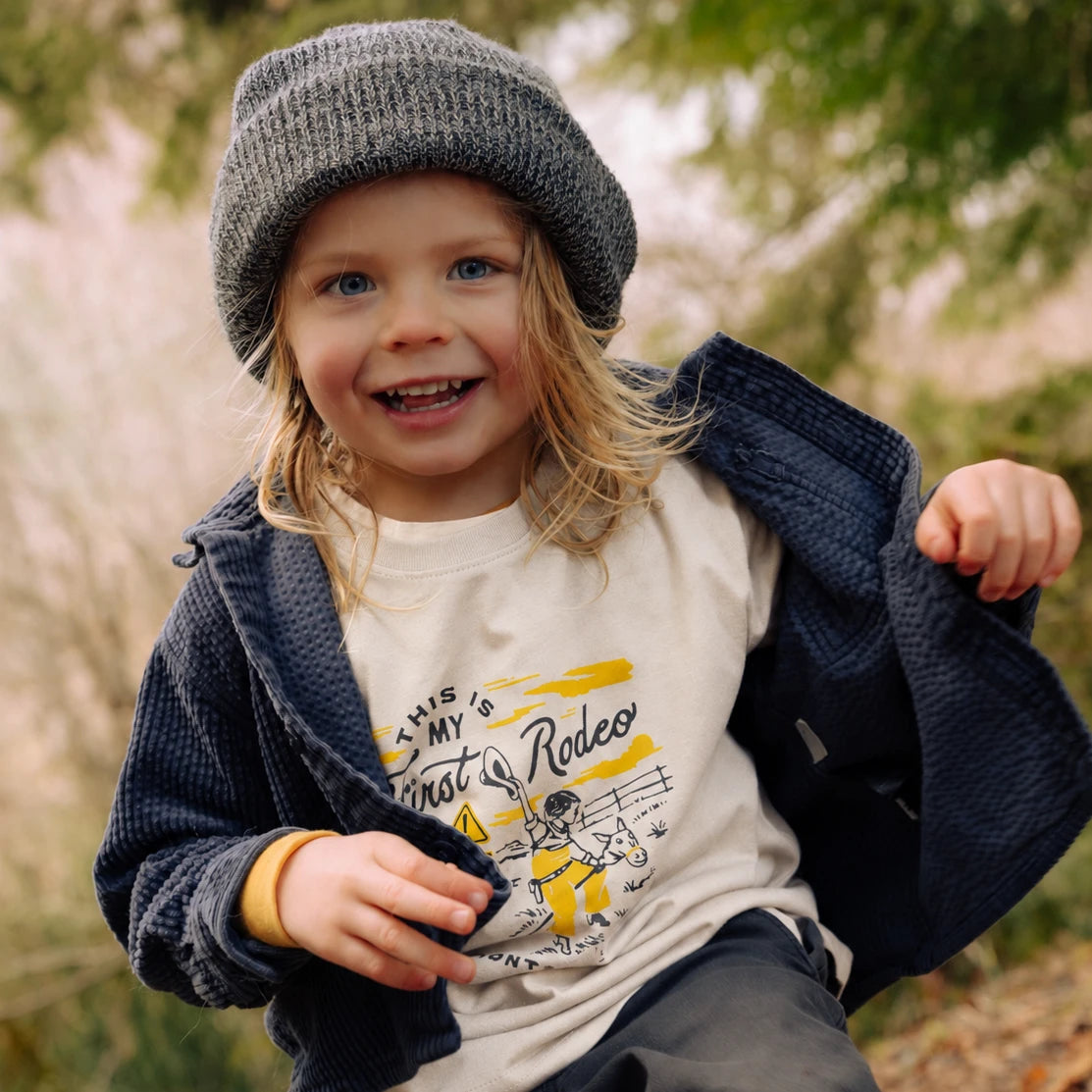 Smiling toddler wearing the natural First Rodeo Toddler tee with a stick horse cowpoke graphic