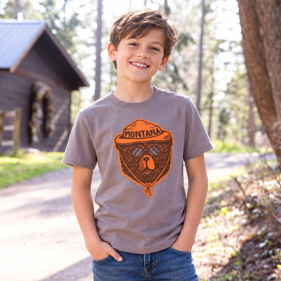 Smiling kid wearing the heather Montana Bear Ranger youth tee with an orange bear ranger graphic