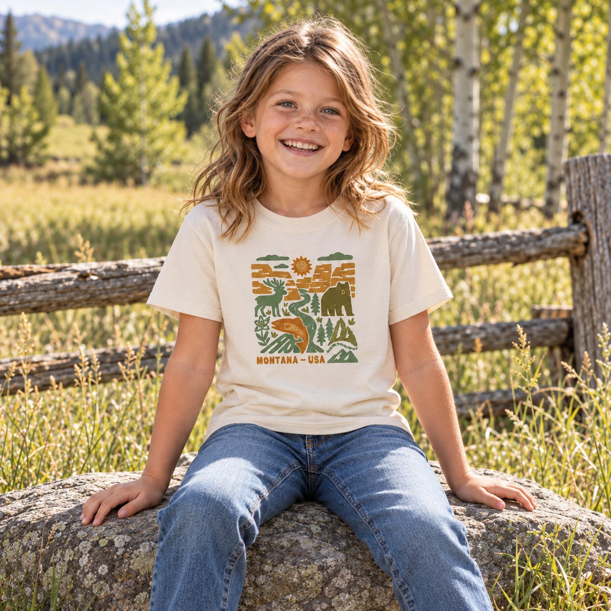 Smiling kid sitting on a rock wearing the natural Blockprint Backcountry kids tee with a bear, buck and trout graphic