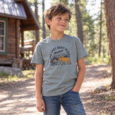 Smiling kid by a log cabin wearing the green Last Best Place Moose kids tee with a bull moose and campfire graphic