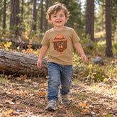 Smiling toddler walking through the forest in the tan Montana Bear Ranger toddler tee